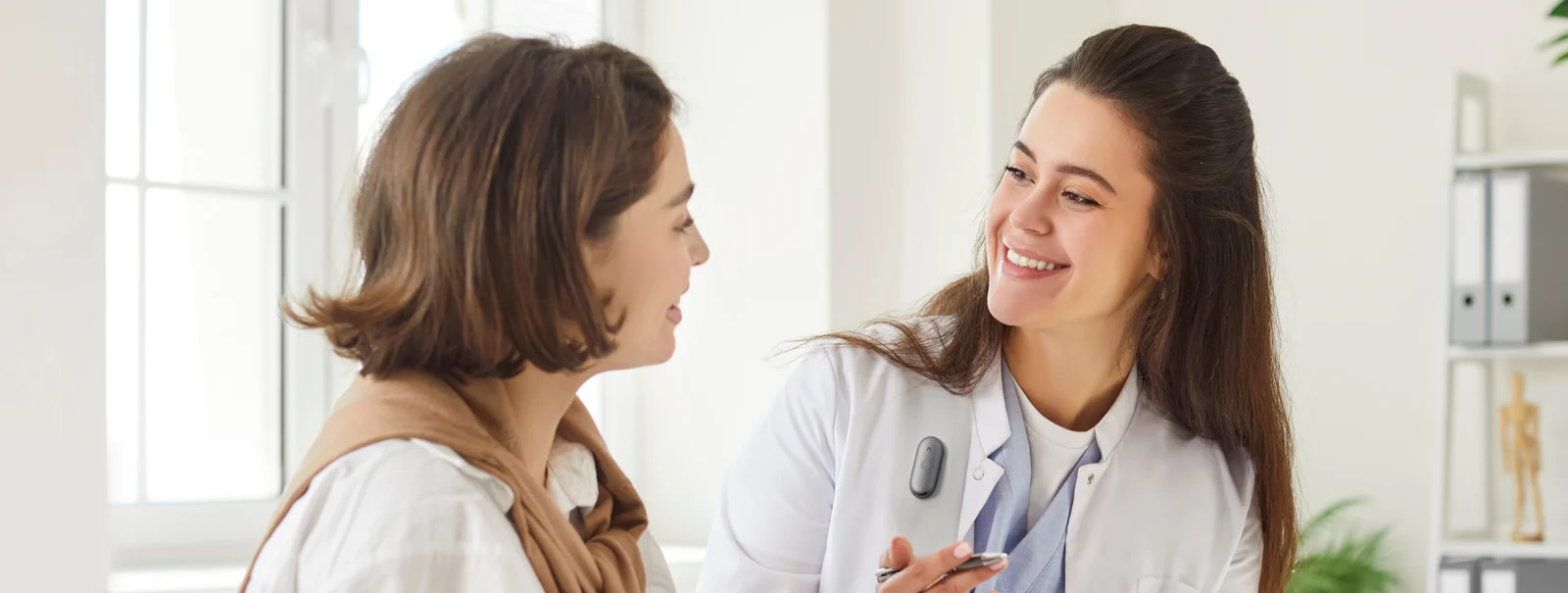 A smiling doctor wearing a Plaud recorder pinned to her white coat while she talks with a patient in a bright exam room.